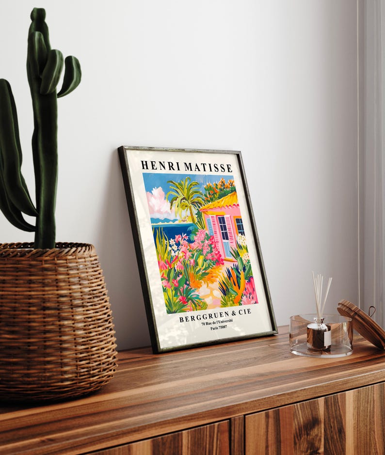 May include: A framed print of a painting by Henri Matisse titled "The Pink House". The painting depicts a pink house with a blue door and a lush garden with palm trees and flowers. The print is framed in black and has the artist's name and the gallery's name and address at the bottom.