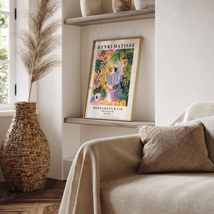 May include: A framed Henri Matisse art print with a gold frame, featuring a colorful illustration of figures reading in a garden setting. The print is displayed on a shelf above a sofa with a tan throw and a decorative pillow. A large woven vase with dried plants is in the foreground.
