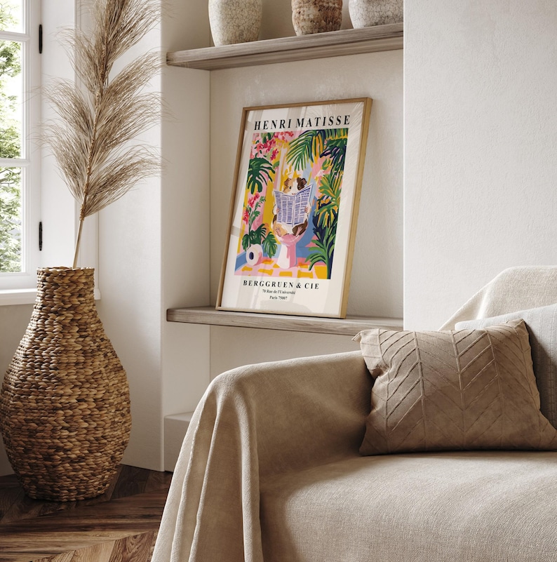 May include: A framed Henri Matisse art print with a gold frame, featuring a colorful illustration of figures reading in a garden setting. The print is displayed on a shelf above a sofa with a tan throw and a decorative pillow. A large woven vase with dried plants is in the foreground.