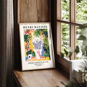 May include: A framed art print displaying a white dog reading a newspaper, set against a vibrant background of tropical plants and flowers. The print is labelled "HENRI MATISSE" and "BERGGRUEN & CIE" in a wooden frame.