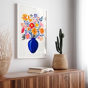 Op de afbeelding: Een wit ingelijste print van een kleurrijk bloemenboeket in een blauwe vaas. De bloemen zijn geschilderd in tinten rood, oranje, geel, roze en groen. De vaas heeft een eenvoudige, ronde vorm.