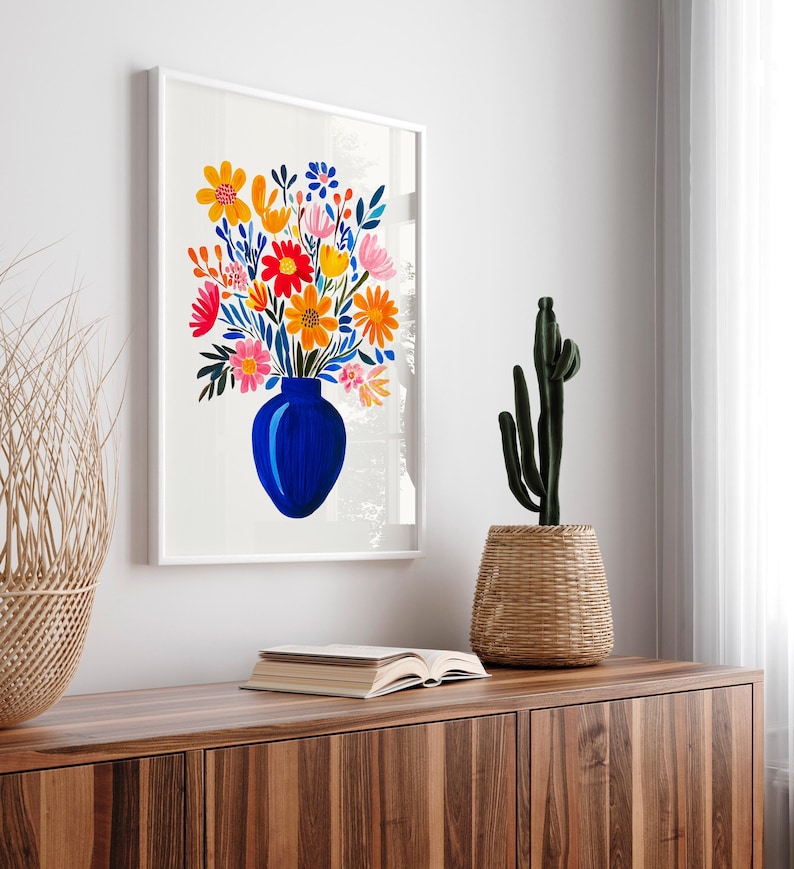 Op de afbeelding: Een wit ingelijste print van een kleurrijk bloemenboeket in een blauwe vaas. De bloemen zijn geschilderd in tinten rood, oranje, geel, roze en groen. De vaas heeft een eenvoudige, ronde vorm.