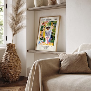 May include: A framed art print featuring a dog reading a newspaper, with the text "HENRI MATISSE" and "BERGGRUEN & CIE". The artwork has a colorful, botanical background with a gold frame. A woven vase with dried plants is in the foreground.