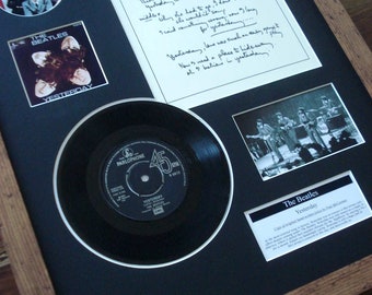 May include: A framed display featuring a black vinyl record of The Beatles' song "Yesterday." The record is surrounded by a white mat and a black background. The frame is made of wood with a brown finish. The display also includes a handwritten lyric sheet for the song, a small photo of the band, and a small photo of the band's album cover.