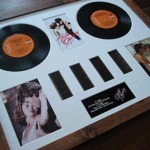 May include: A framed display featuring a 45 rpm record, film strips, and photos from the movie "Dirty Dancing". The record is black with an orange label and the title "The Time Of My Life" is visible. The film strips are black and white and the photos are in colour. The frame is brown wood.