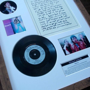 Op de afbeelding: Een ingelijste weergave van een 45 RPM vinylplaat van Michael Jacksons nummer "Beat It". De weergave bevat een zwart-witfoto van Michael Jackson, een handgeschreven tekstblad en een kleinere foto van Michael Jackson in een rood jack. De tekst op het tekstblad luidt "Beat It" en bevat songteksten uit het nummer.
