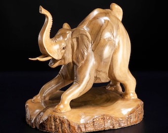 May include: A wooden sculpture of an elephant in a crouching pose. The elephant is carved from a light brown wood with a natural wood grain. The sculpture is set on a wooden base.