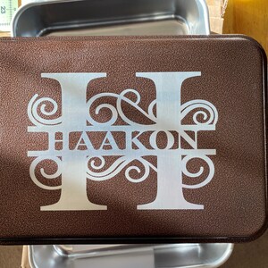 May include: A brown metal baking pan with a white monogram design featuring the letters "HH" and the name "HAAKON" in a decorative font.