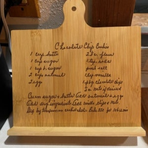 May include: A wooden cutting board with a recipe for chocolate chip cookies handwritten in brown ink. The recipe includes ingredients like butter, eggs, flour, sugar, chocolate chips, and vanilla. The board has a handle and a stand.