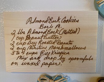 May include: A handwritten recipe for Almond Bark Cookies on a wooden surface. The recipe includes 2 pounds of almond bark, 1 cup of peanut butter, 2 cups of dry roasted peanuts, 2 cups of miniature marshmallows, and 3 to 4 cups of Rice Krispies. The recipe instructs to mix the ingredients and drop by spoonfuls on waxed paper.