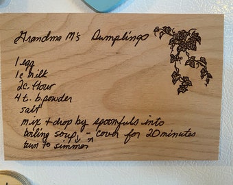 May include: A wooden recipe card with handwritten instructions for making Grandma M's Dumplings. The recipe calls for 1 egg, 1 cup of milk, 2 cups of flour, 4 teaspoons of baking powder, and salt. The instructions are to mix and drop by spoonfuls into boiling soup, cover for 20 minutes, and then turn to simmer.