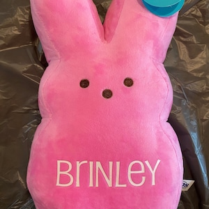 May include: Pink Peeps bunny plush toy with the name "Brinley" embroidered on the front. The toy has a brown nose and eyes and a blue "Peeps" tag attached.