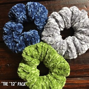 May include: Three scrunchies in blue, gray, and green velvet. The scrunchies are arranged in a circle on a wooden surface. The text "THE "12" PACK" is visible at the bottom of the image.