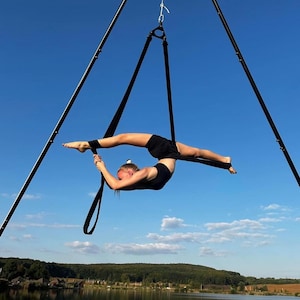 May include: A person performing aerial yoga, suspended from a black metal rig against a blue sky. The person is wearing black athletic wear and is in a split position, holding onto the black straps. A lake and trees are visible in the background.