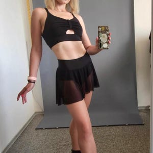 Pode incluir: Uma mulher vestindo um sutiã esportivo preto com um detalhe de corte e uma saia de malha preta. Ela está usando botas de salto alto pretas e segurando um smartphone na mão direita.