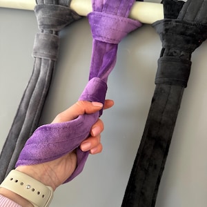 May include: Three velvet yoga swing straps in gray, purple, and black are suspended from a white bar. The purple strap is held by a hand. The straps are designed for aerial yoga and fitness.