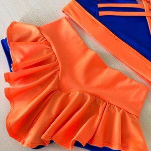 May include: A two-piece dance outfit with a bright orange skirt and a blue top. The skirt has a ruffled hem and the top has a criss-cross back design.