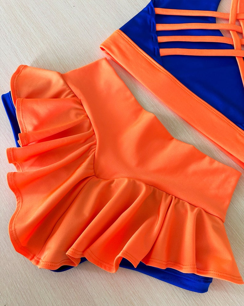 May include: A two-piece dance outfit with a bright orange skirt and a blue top. The skirt has a ruffled hem and the top has a criss-cross back design.