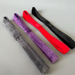 Op de afbeelding: Vier haarbandjes van fluweel in verschillende kleuren: grijs, paars, rood en zwart. De bandjes zijn gevouwen en in een strik geknoopt.