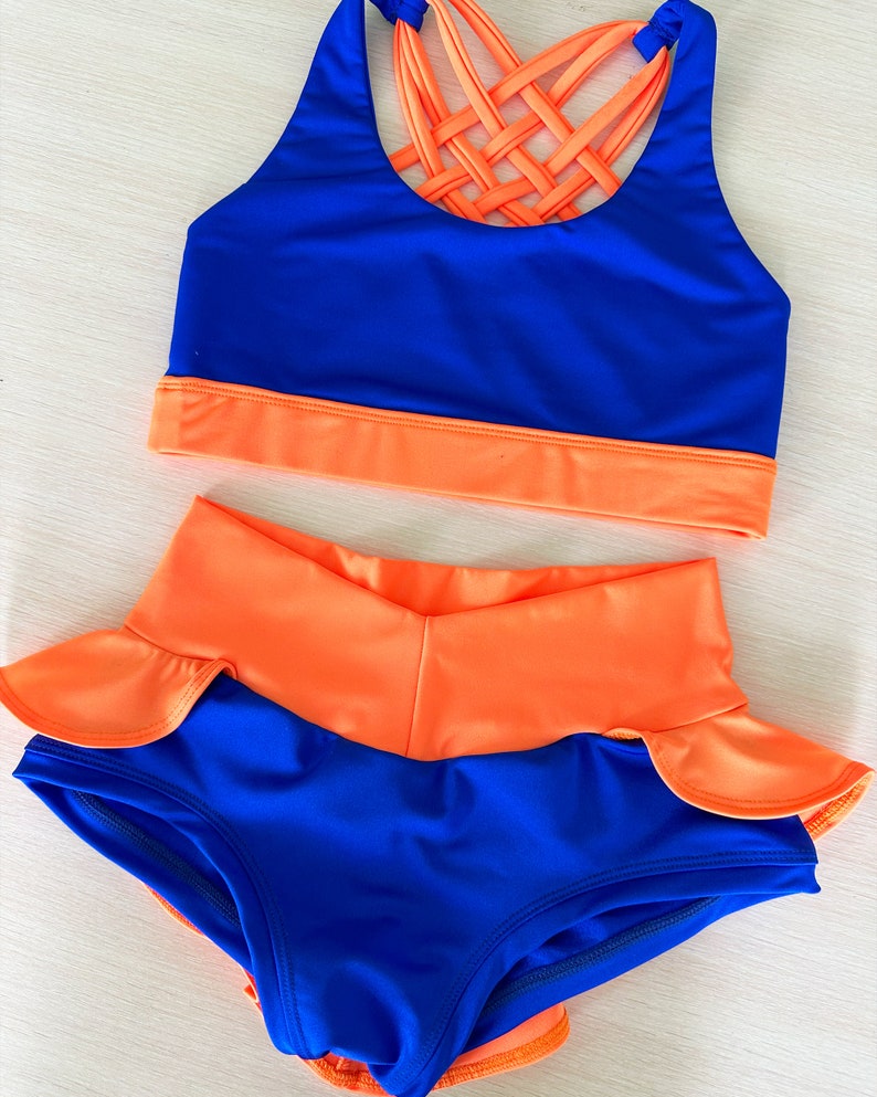 May include: A two-piece dance outfit with a blue sports bra and orange trim. The bra has a criss-cross design at the back. The matching shorts are orange with a blue ruffle trim.