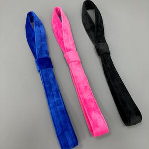 May include: Three velvet headbands in vibrant colors: royal blue, hot pink, and black. Each headband features a wide, ruched band and a loop design. The headbands are displayed against a neutral gray background.