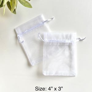 May include: Two white sheer organza gift bags with white ribbon ties. The bags are 4 inches by 3 inches.