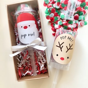 May include: A Christmas-themed confetti popper set. One popper features a Santa Claus design with the text "pop me!" and is packaged in a brown box with red and white crinkle paper. The other popper has a reindeer design and the text "POP ME!"