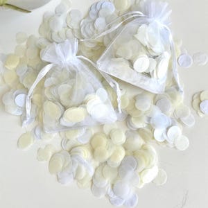 May include: Two white organza bags filled with white and ivory paper confetti. The bags are on a white surface with loose confetti scattered around them.