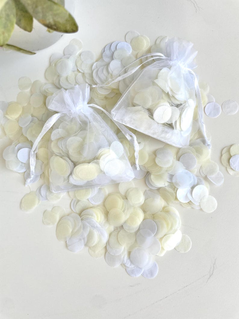 May include: Two white organza bags filled with white and ivory paper confetti. The bags are on a white surface with loose confetti scattered around them.