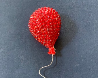 Puede incluir: Un pin de globo rojo de pedrería con una cola de alambre plateado.