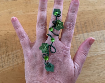 Puede incluir: Un anillo de vid verde con hojas y gemas verdes. El anillo se lleva en un dedo.