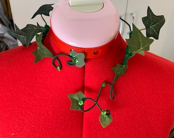 Puede incluir: Un collar de hiedra verde con hojas verdes y pequeñas cuentas verdes. El collar se muestra en un maniquí rojo.