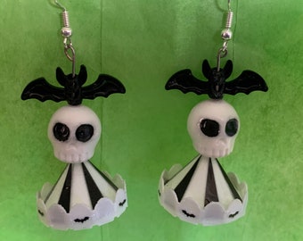 Puede incluir: Un par de pendientes de temática de Halloween con una calavera blanca con ojos negros y un murciélago negro en la parte superior. La calavera está unida a un cono a rayas blanco y negro con un borde festoneado y pequeños murciélagos alrededor de la parte inferior.
