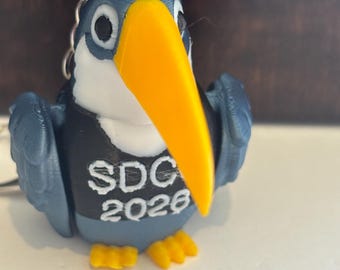Puede incluir: Un llavero de pájaro impreso en 3D con cuerpo azul, pecho blanco y un gran pico amarillo. El pájaro lleva un chaleco negro con el texto blanco SDC 2026 impreso en la parte delantera. Tiene patas pequeñas amarillas y una anilla metálica.