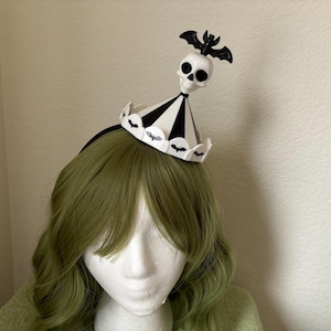 May include: A black and white headband with a skull and bat design. The headband is shaped like a party hat with black and white stripes. The skull is white with black eyes and a black bat is perched on top of the skull.