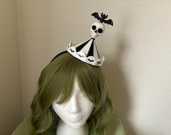 Puede incluir: Una diadema negra y blanca con un diseño de calavera y murciélago. La diadema tiene forma de gorro de fiesta con rayas negras y blancas. La calavera es blanca con ojos negros y un murciélago negro está posado en la parte superior de la calavera.