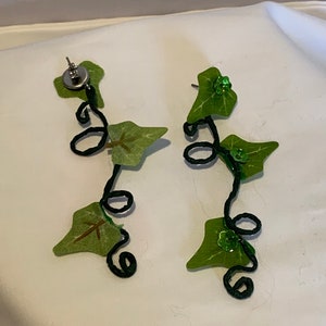 May include: A pair of dangle earrings featuring a black wire vine with green fabric leaves and green glitter accents.