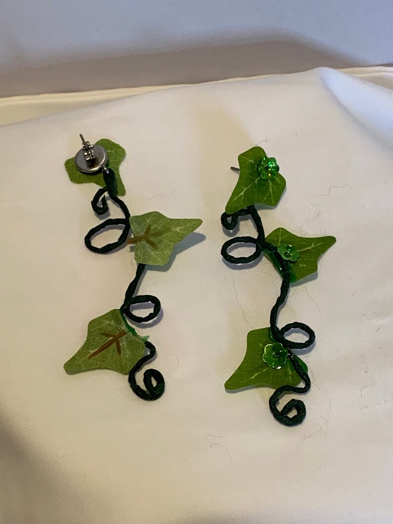 May include: A pair of dangle earrings featuring a black wire vine with green fabric leaves and green glitter accents.