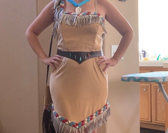 Puede incluir: Una mujer con un vestido marrón con flecos y un collar turquesa. El vestido tiene un cinturón marrón con detalles en turquesa y verde. Lleva un bolso marrón con flecos.