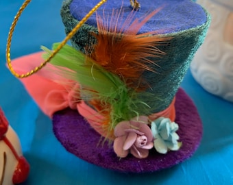 Puede incluir: Un sombrero de copa de fieltro azul y morado con una cinta rosa, plumas verdes y naranjas y dos flores de tela. El sombrero tiene un lazo de cuerda dorado para colgar.