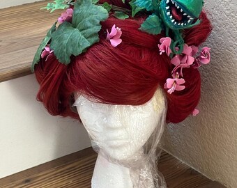 Puede incluir: Una peluca roja adornada con hojas verdes, flores rosas y una venus atrapamoscas verde de dibujos animados. La peluca se exhibe en un maniquí de espuma blanco, envuelta en plástico y colocada sobre una superficie de madera.