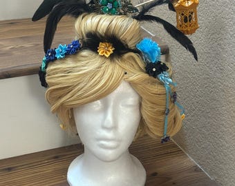 Puede incluir: Peluca rubia adornada con plumas negras, adornos florales y una pequeña linterna decorativa. La peluca se exhibe en una cabeza de maniquí blanca, destacando el diseño detallado. Los elementos florales presentan acentos azules, amarillos y negros.