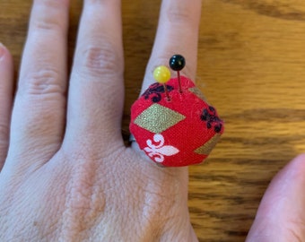 Puede incluir: Un cojín de alfileres de tela roja con detalles dorados y negros, con forma de anillo, con dos alfileres insertados.