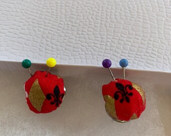 Puede incluir: Dos alfileteros de tela roja con patrones geométricos dorados y negros y un diseño de flor de lis. Cada alfiletero está coronado con un alfiler con una cabeza de plástico de color.