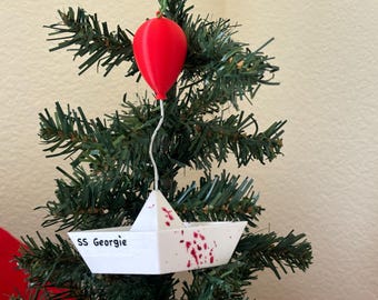 Puede incluir: Un adorno de Navidad de barco de papel blanco con salpicaduras de sangre roja y el texto "SS Georgie" colgado de un árbol de Navidad artificial verde. Un globo rojo está unido a la parte superior del barco.