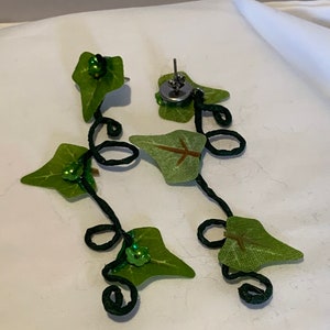 May include: A pair of green leaf earrings with black wire and green sequins. The earrings are in the shape of a vine with leaves and are attached to a silver post.