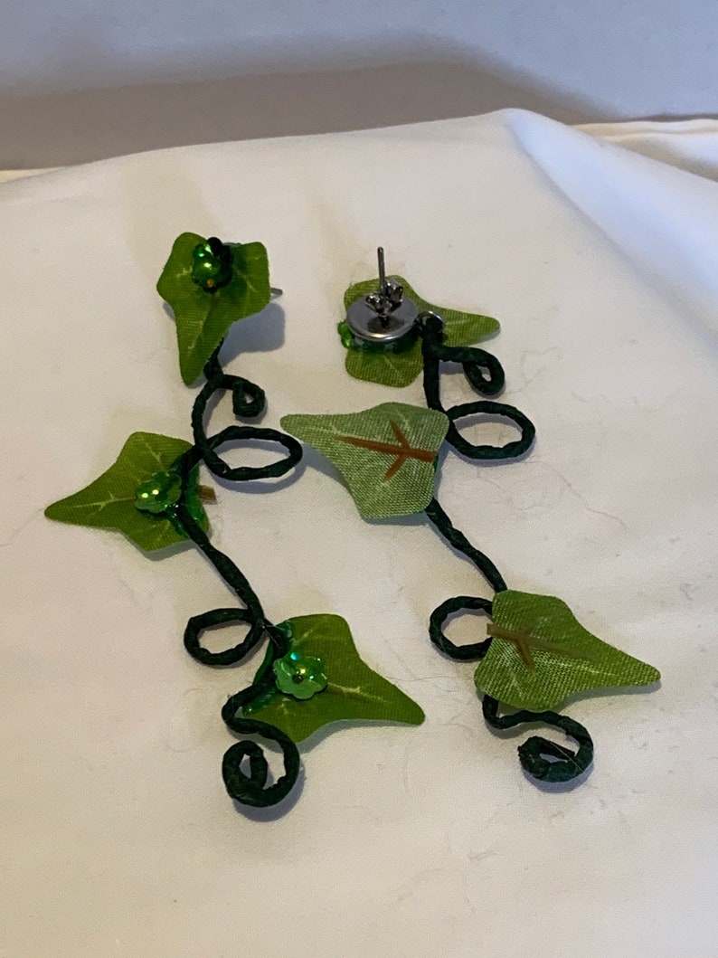 May include: A pair of green leaf earrings with black wire and green sequins. The earrings are in the shape of a vine with leaves and are attached to a silver post.