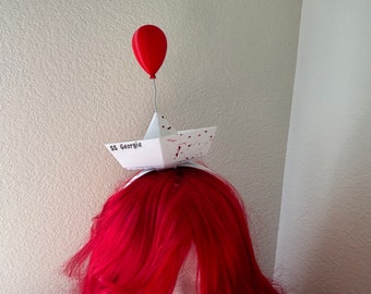 Puede incluir: Un barco de papel blanco con salpicaduras de sangre roja y el texto "SS Georgie" en el lateral. Un globo rojo está sujeto a la parte superior del barco.