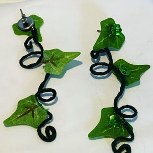 May include: A pair of dangle earrings featuring a black wire vine with green leaves and green glitter accents.