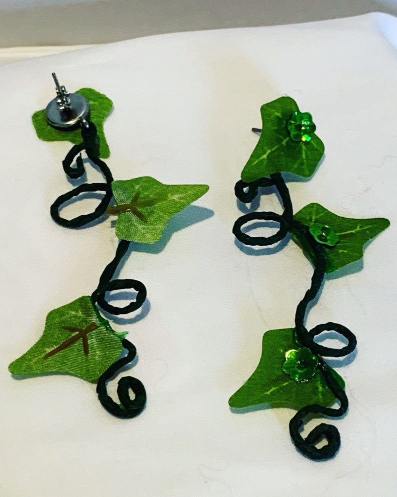 May include: A pair of dangle earrings featuring a black wire vine with green leaves and green glitter accents.